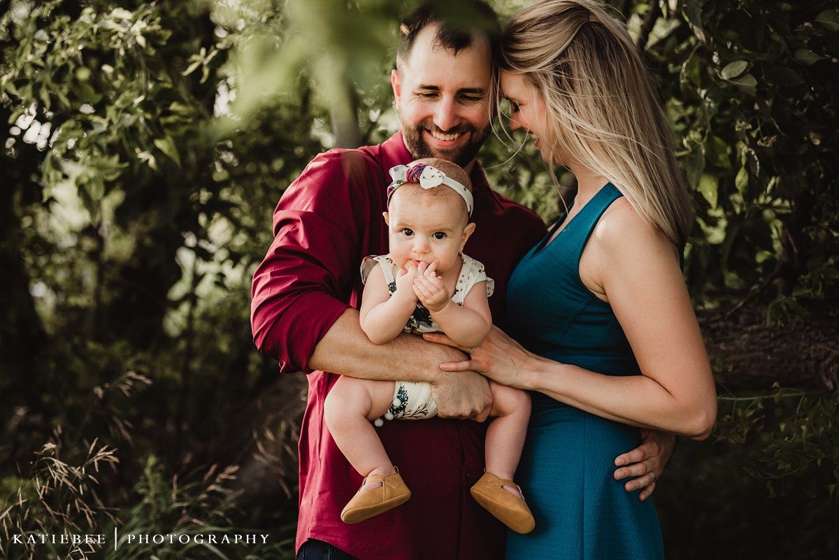 A tender family portrait shows a couple in burgundy and teal embracing their baby outdoors among greenery.