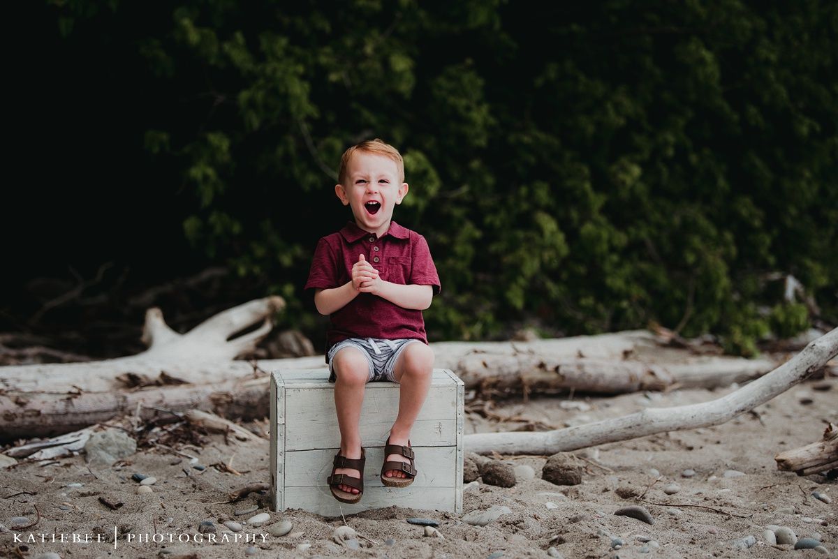 A child sits on a white cube outdoors against a natural backdrop of trees and driftwood.