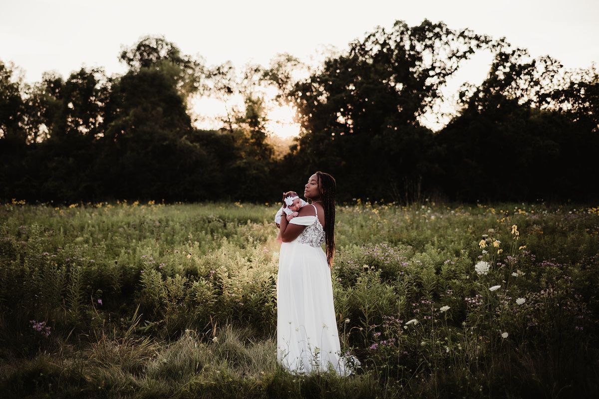 A woman in a flowing white dress holds a baby in a sunset-lit meadow.