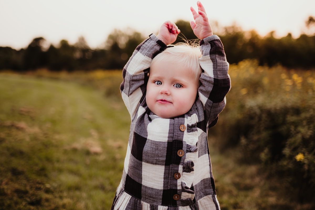 Small child in plaid dress raises arms while playing outdoors at golden hour.