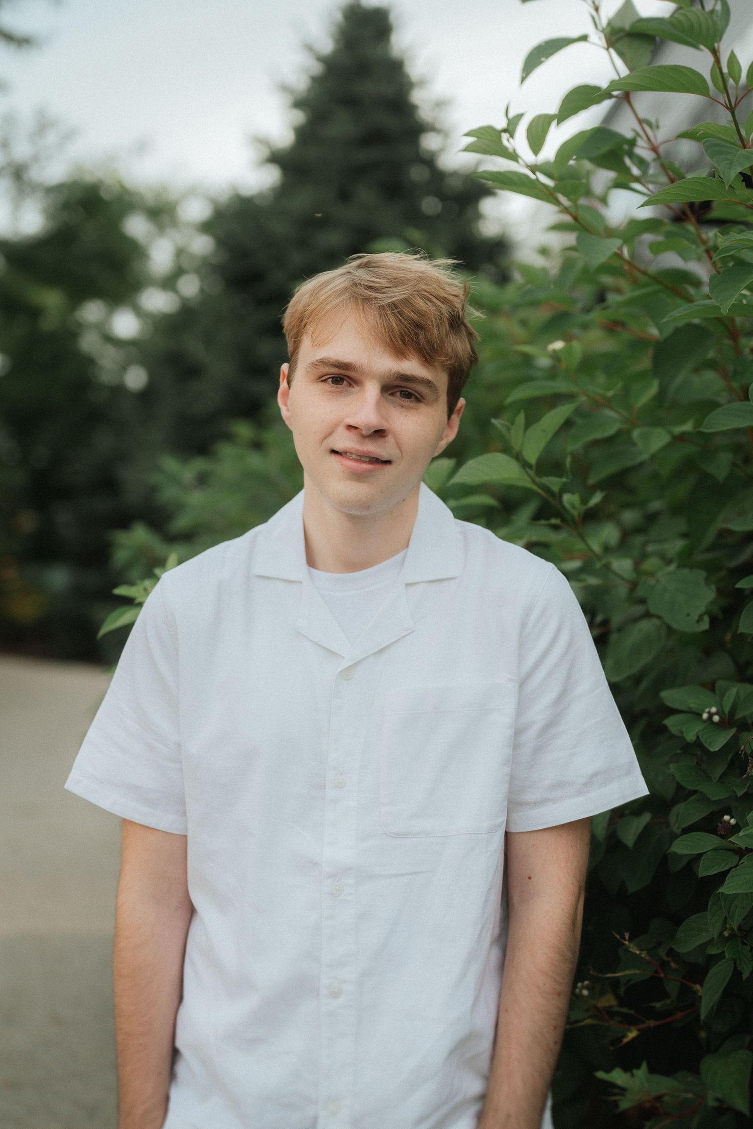 A person wearing a white button-down shirt poses outdoors near leafy plants.