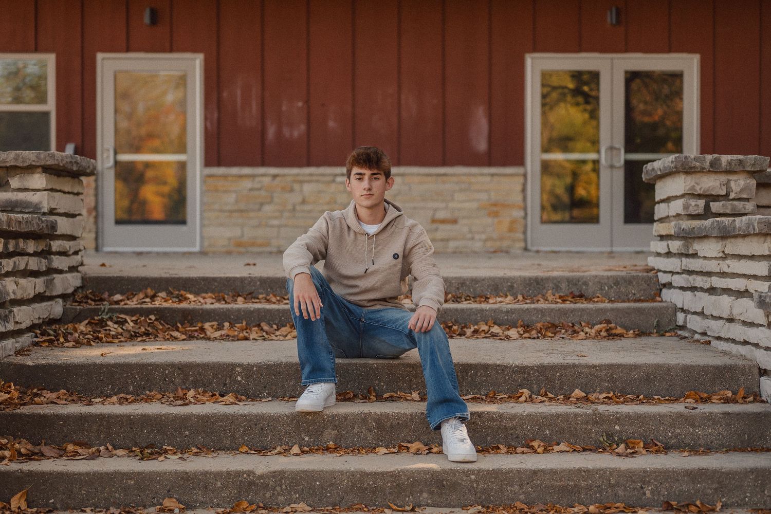 A person sits casually on stone steps outside a brown building surrounded by autumn leaves.