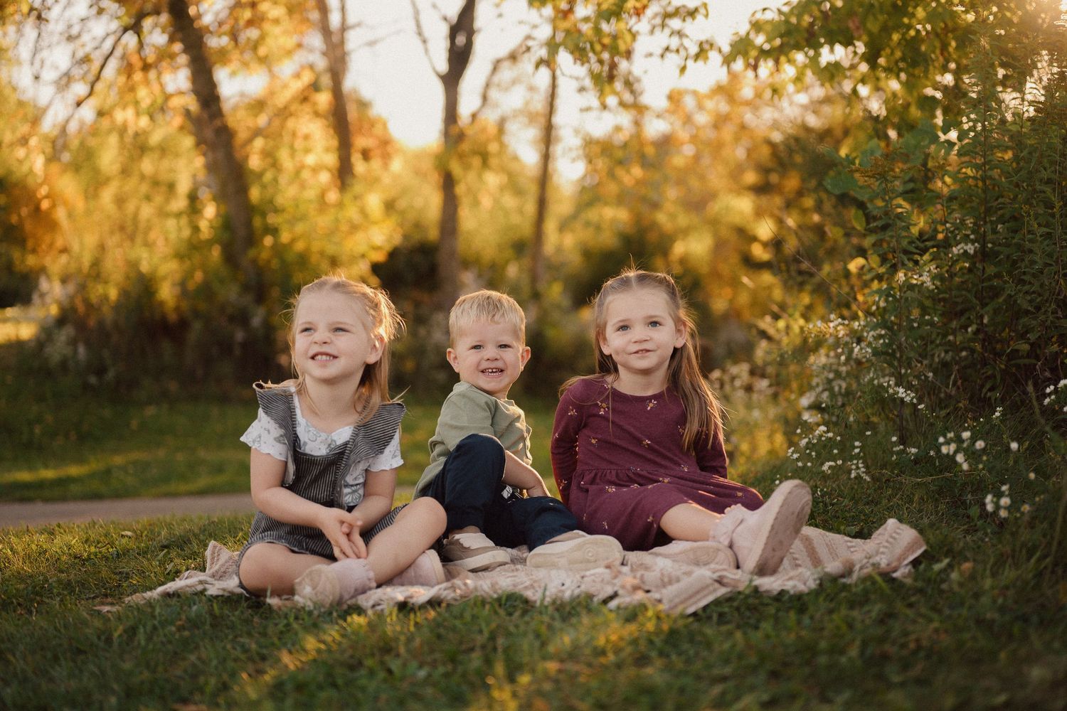Three children sit together on a blanket in a park during golden hour with autumn trees in the background.