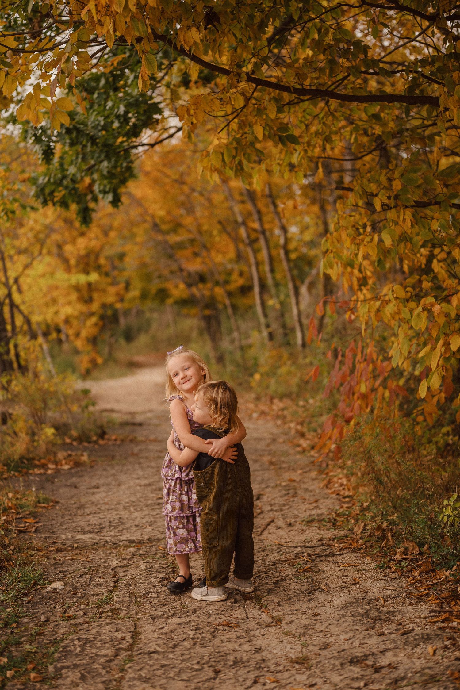 Two children sharing a tender moment on a leaf-covered autumn path.