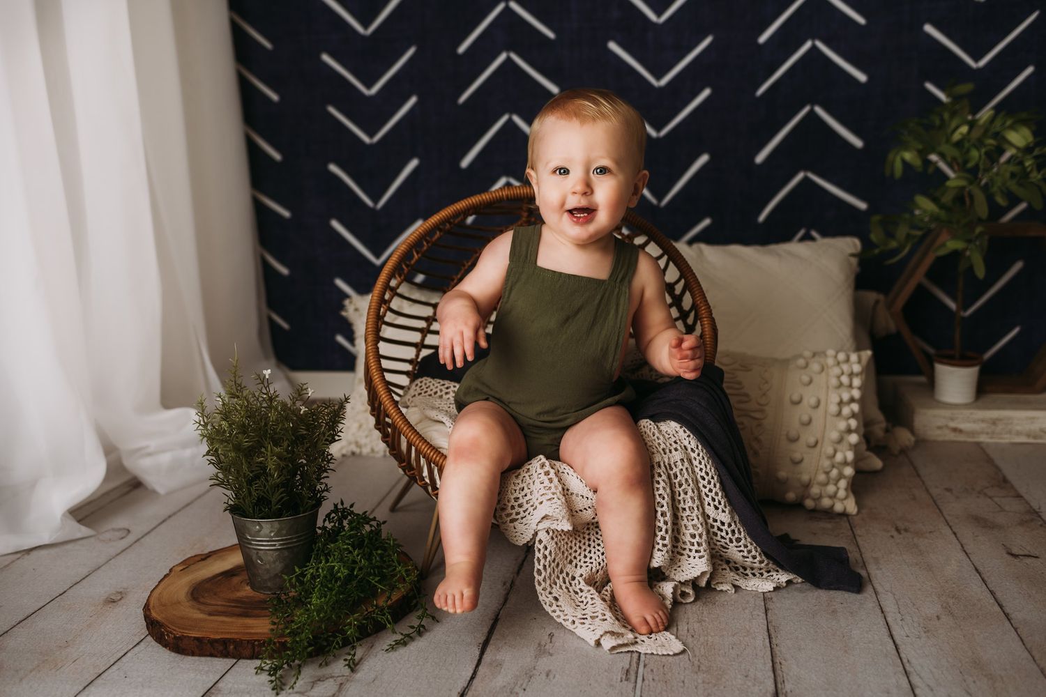 A staged portrait setup featuring a wicker chair against a navy geometric wall with decorative greenery and blankets.