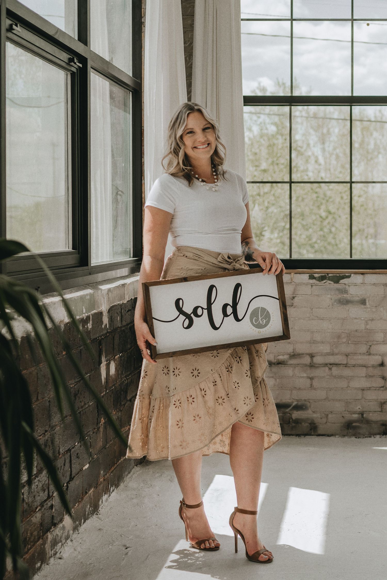 Woman in white top and flowy skirt holds wooden 'sold' sign by industrial windows.