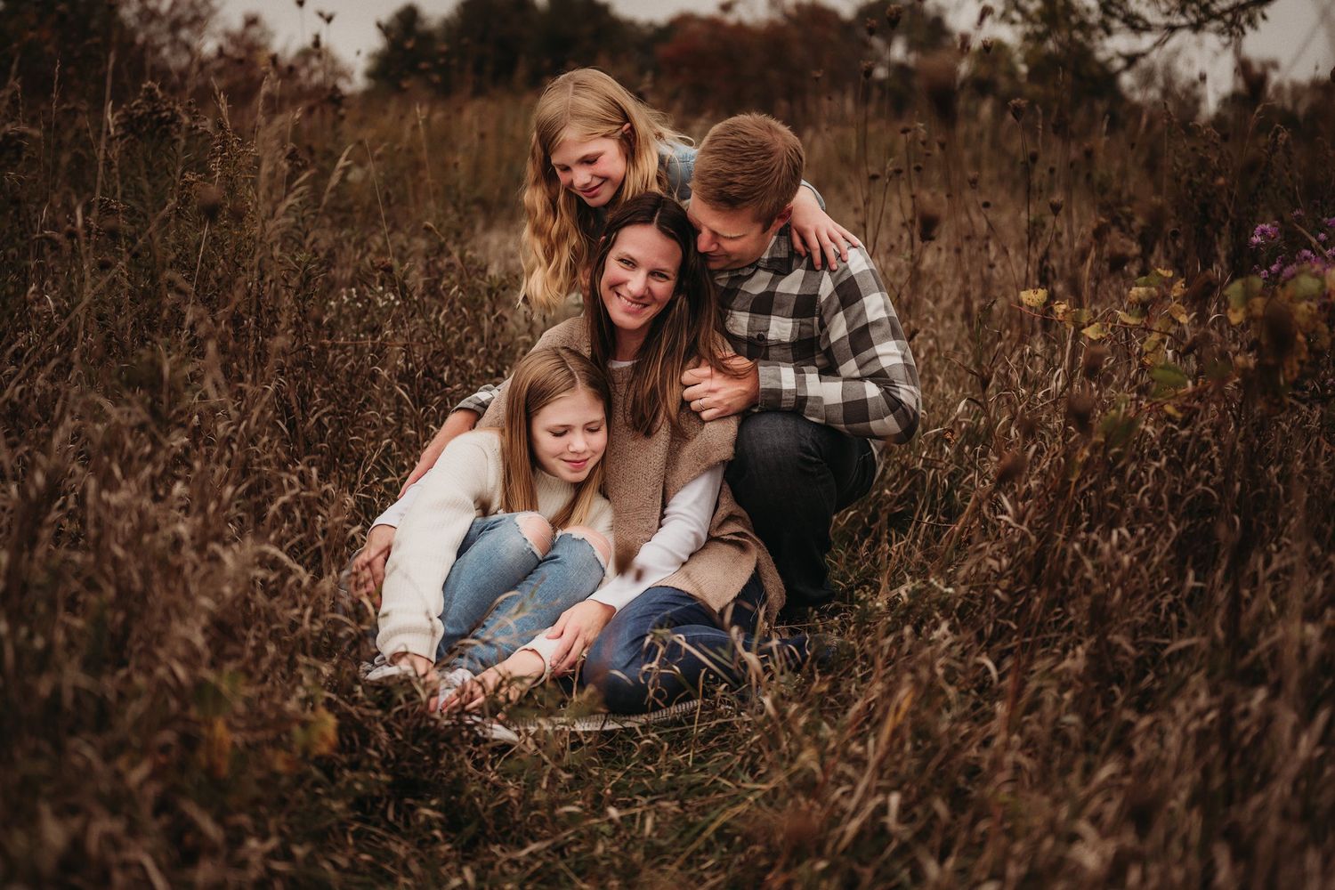 Family of four cuddles together while sitting in autumn field during golden hour.