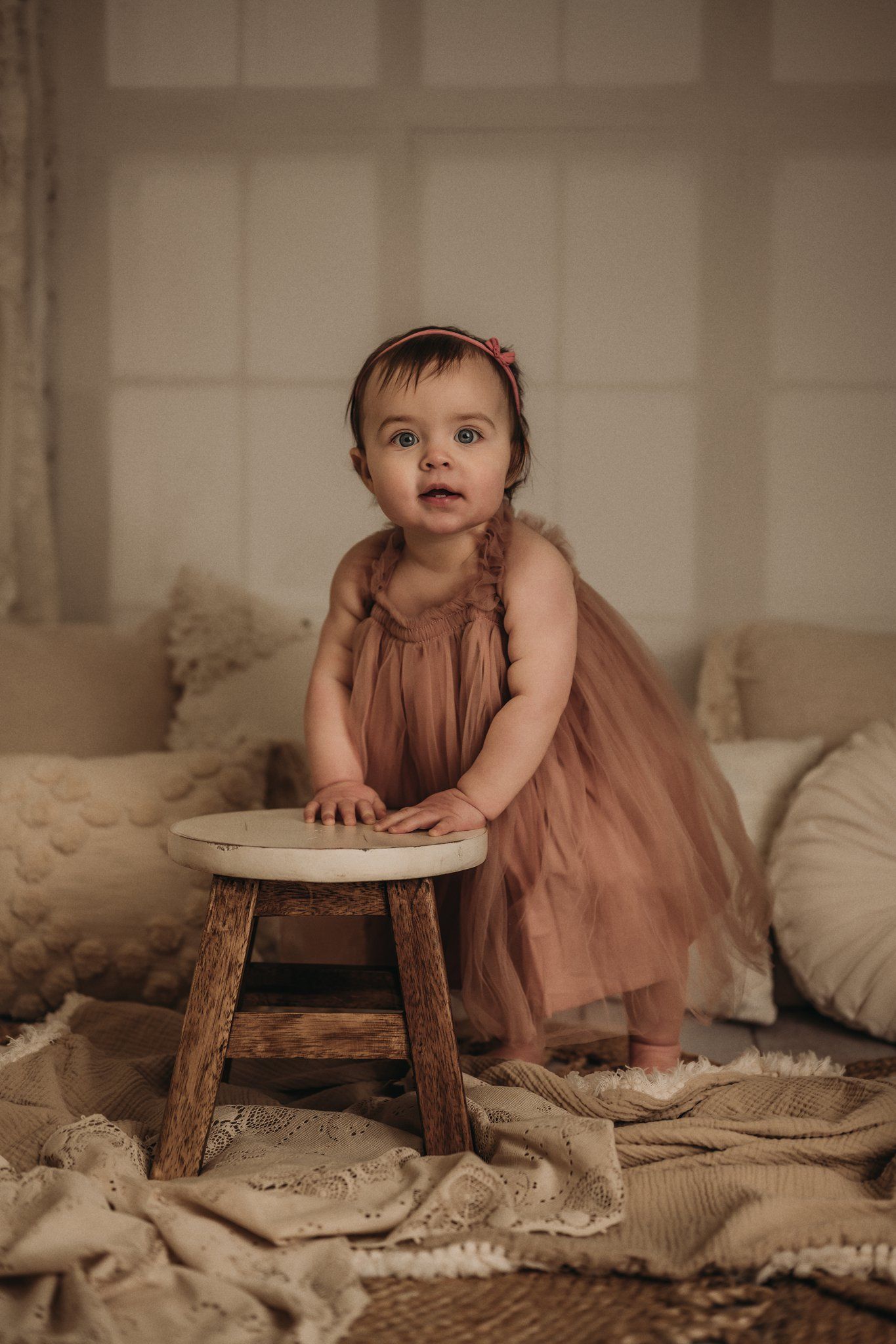 Baby in pink tulle dress stands near rustic wooden stool in cozy room setting.
