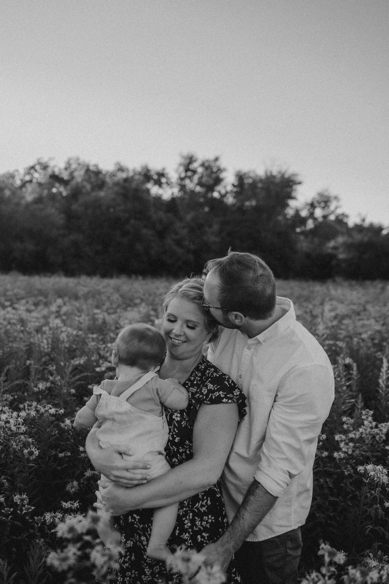 Black and white portrait of a family sharing tender moment in flowering meadow surrounded by trees.