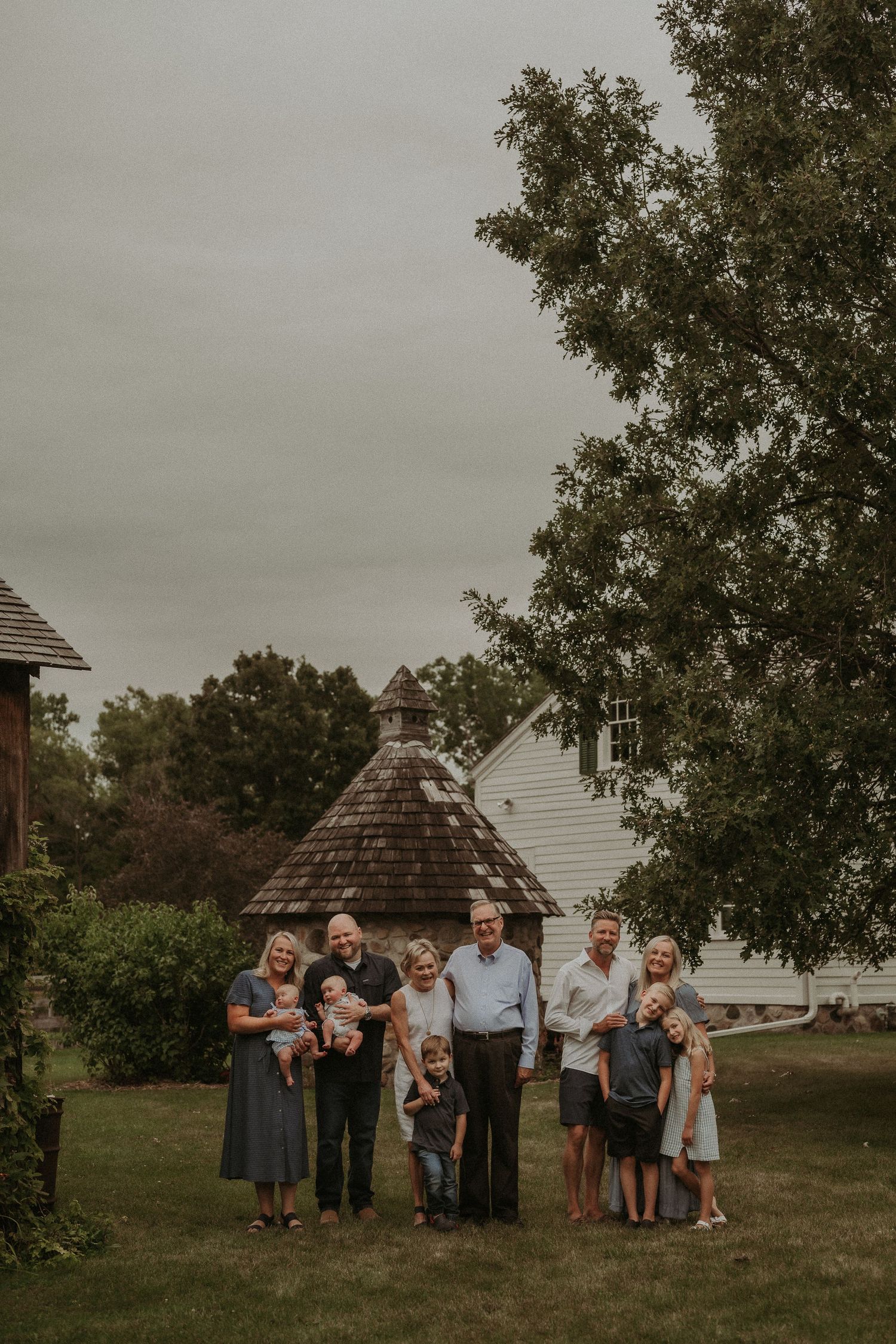 Multi-generational group gathers for outdoor family portrait by rustic white farmhouse on overcast day.