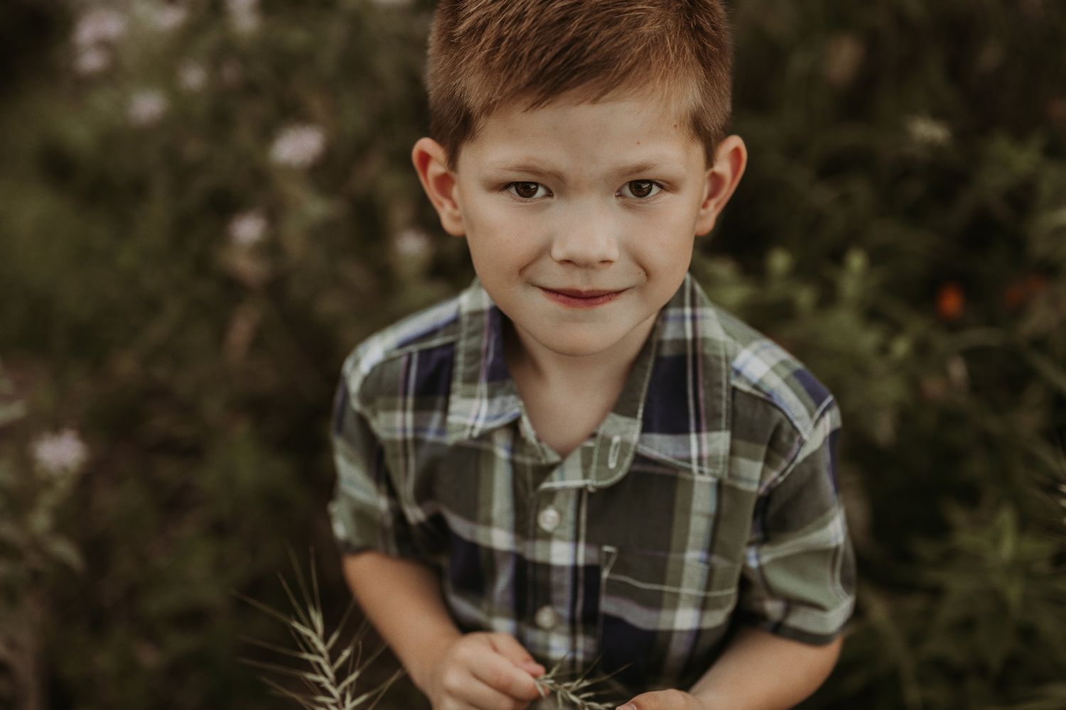 Child wearing plaid button-up shirt smiles warmly while posing in meadow with white flowers.