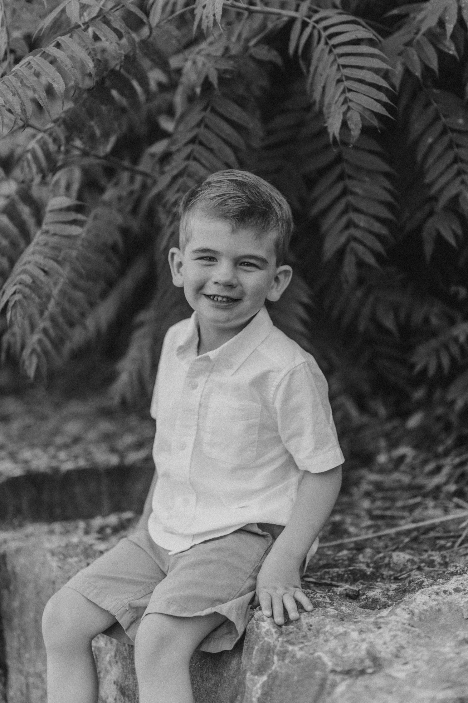 Black and white portrait of child smiling while sitting on stone wall with ferns behind.