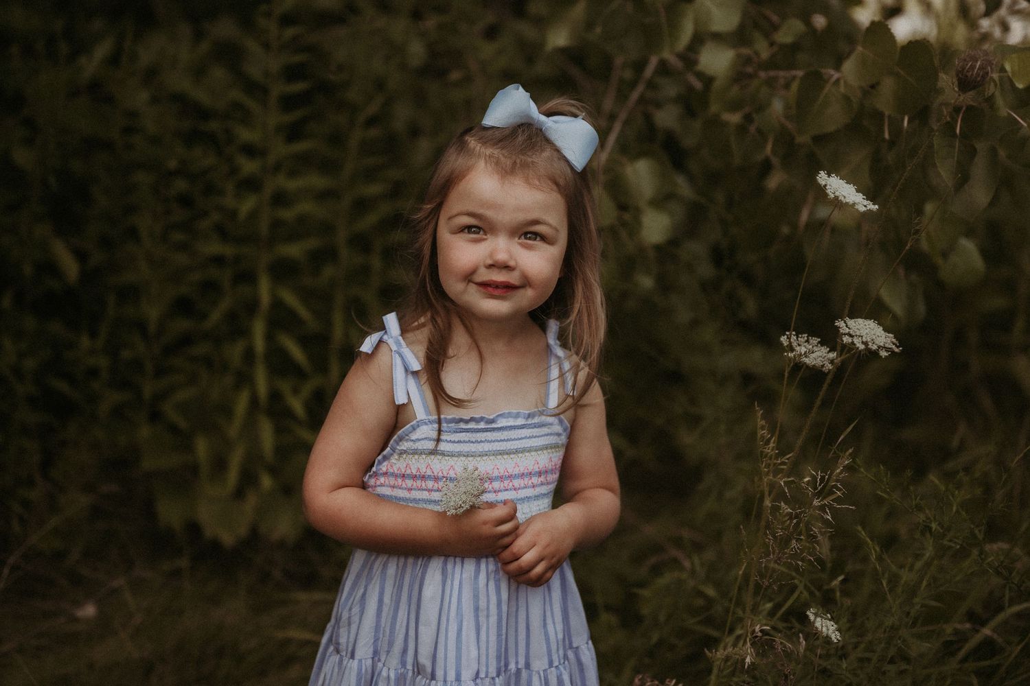 A child in a blue dress with bow accessory poses against a backdrop of wild plants and flowers.