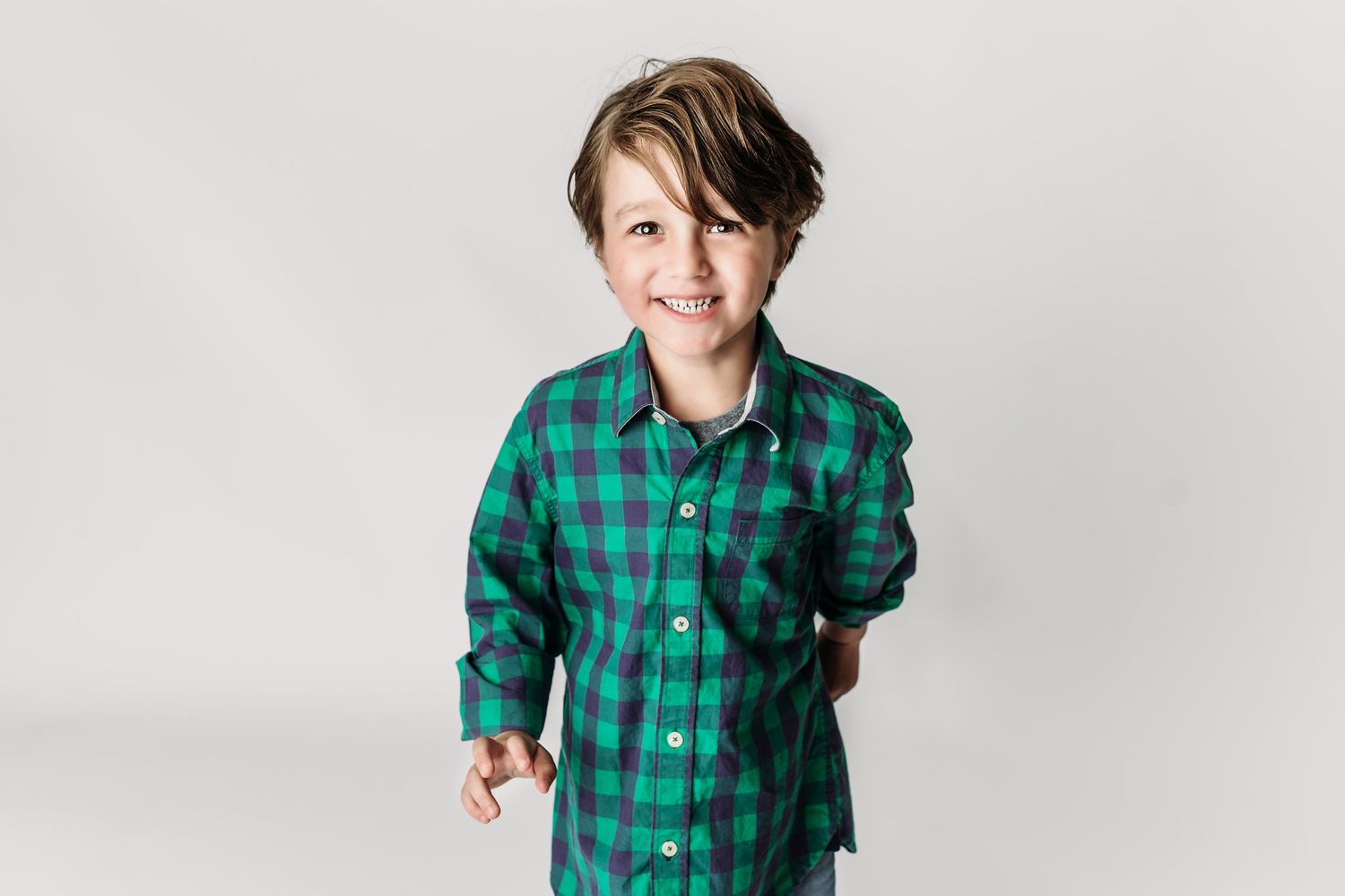 Child in green buffalo check shirt poses playfully against white backdrop.
