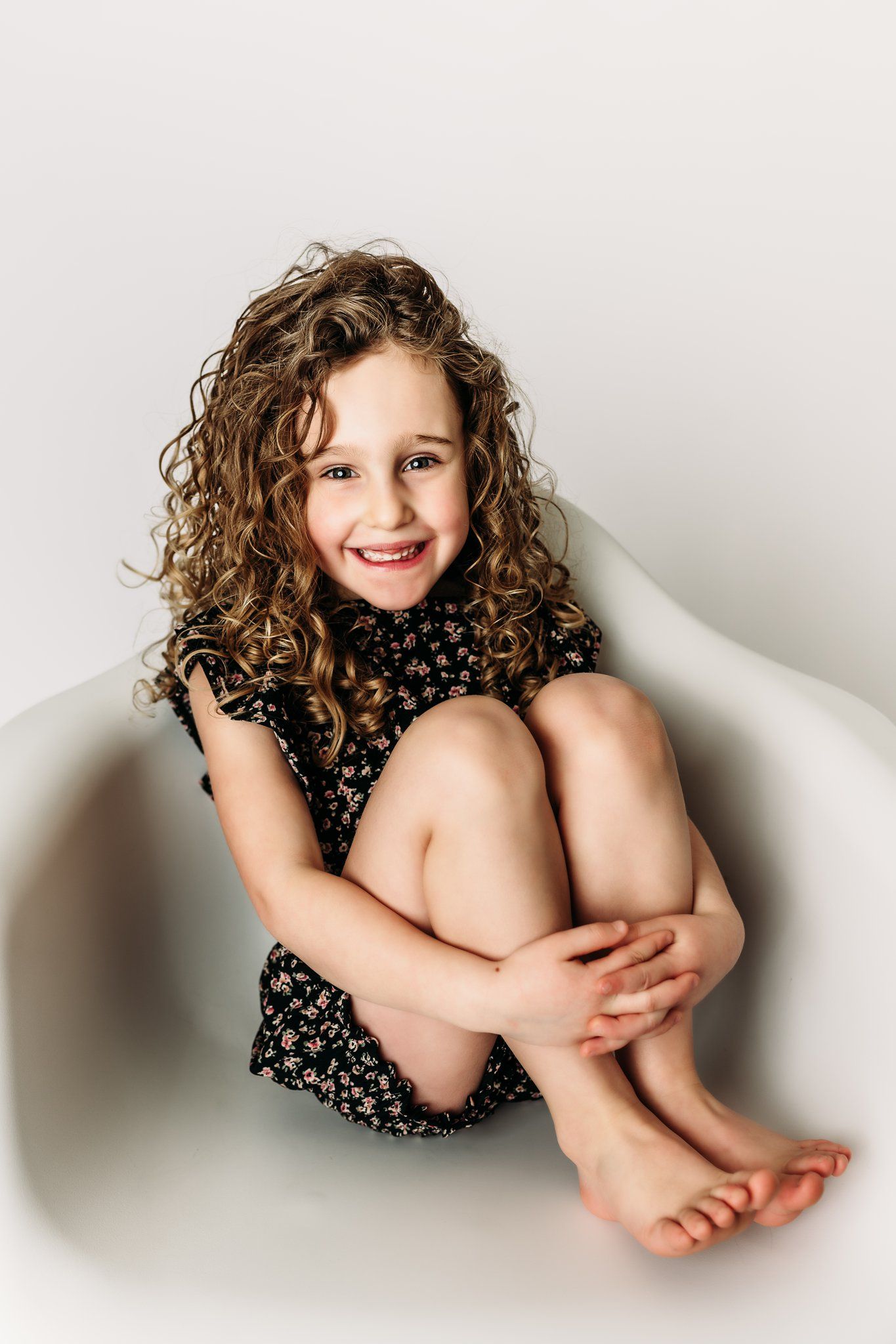 A child with curly hair sits casually in a white chair wearing a floral print dress.