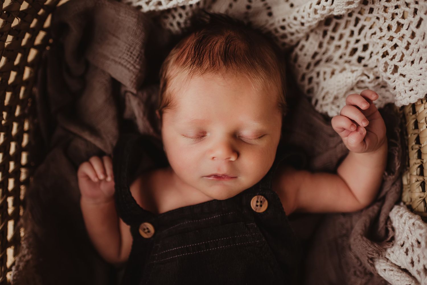 Sleeping baby wearing dark overalls rests peacefully on cream-colored knit blankets.