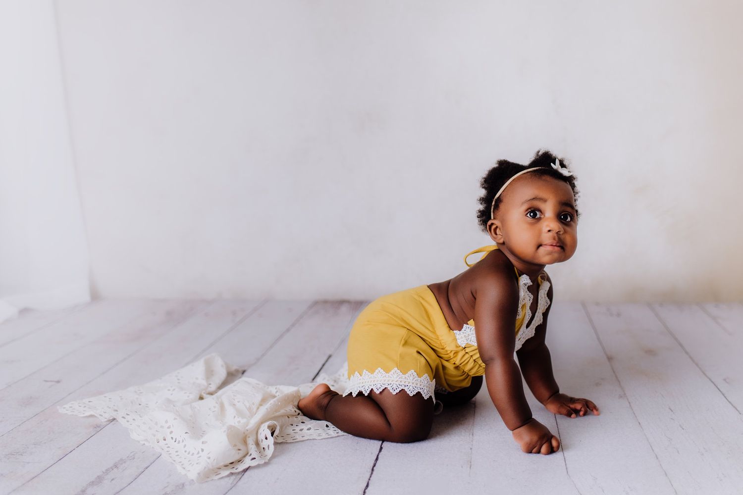 Baby in yellow romper exploring white wooden floor during portrait session.