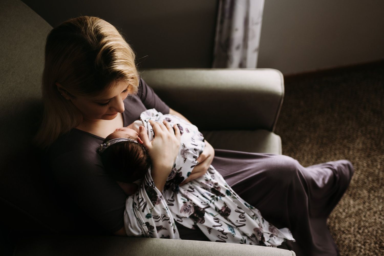 Intimate moment captured of baby being held in grey armchair with natural window light.