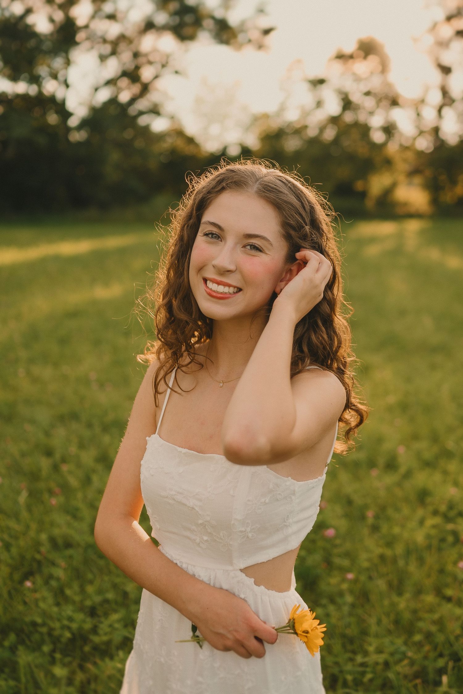 A golden hour portrait in a field shows a person in a white dress holding yellow flowers and smiling warmly.