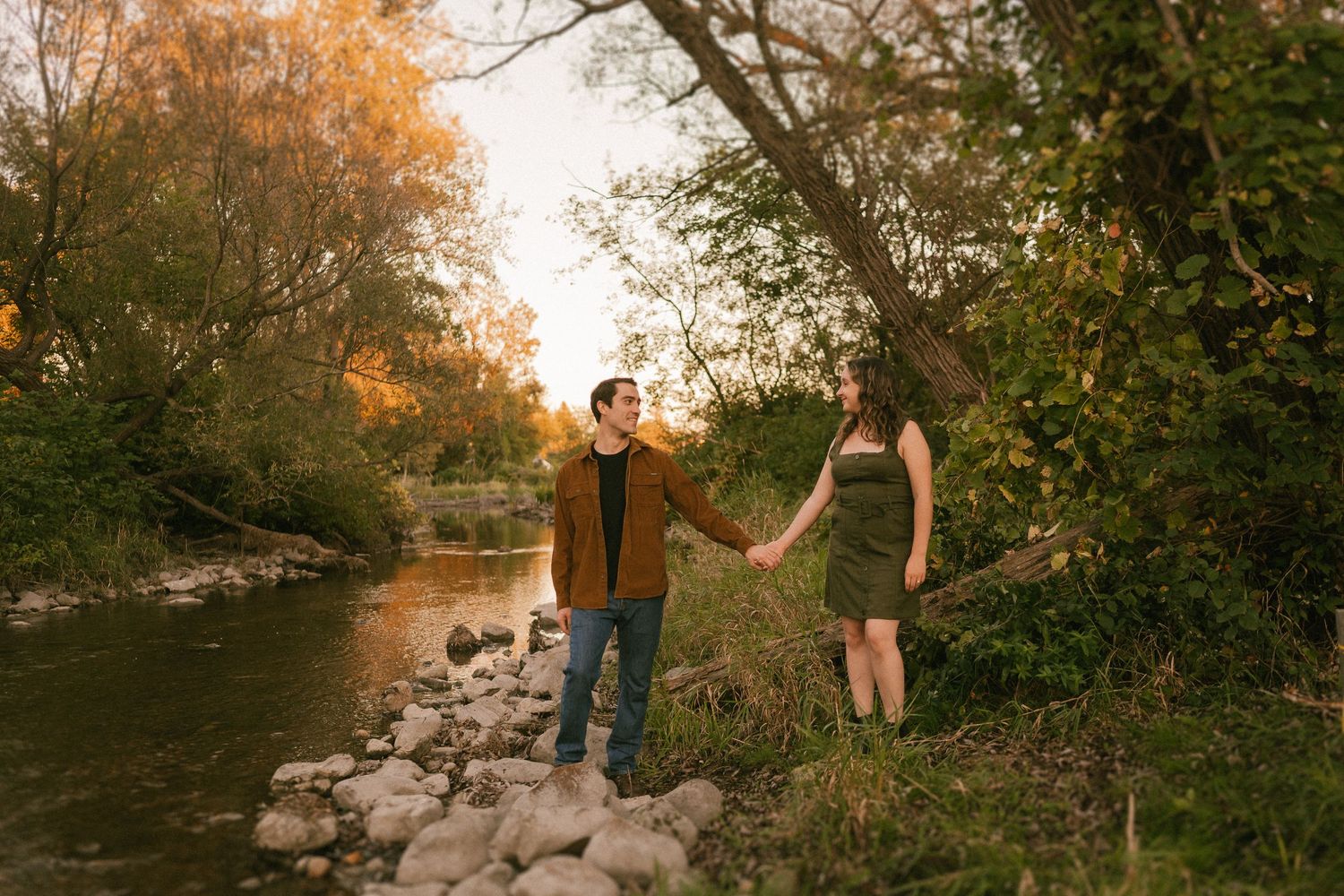 A couple holds hands while standing on rocky riverbank during golden hour surrounded by autumn trees.