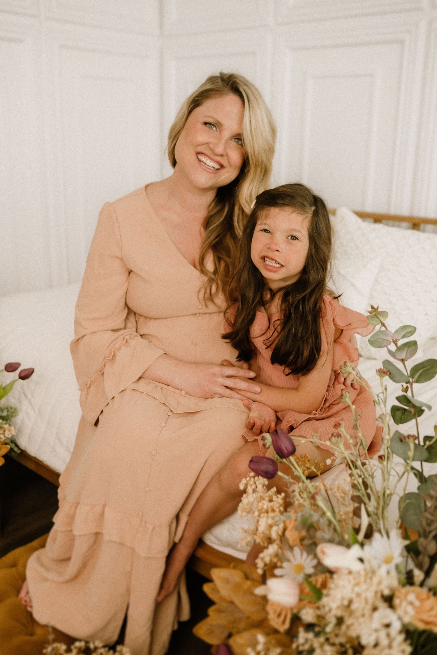 Mother and daughter in matching peach dresses pose together among floral decorations in a bright white room.