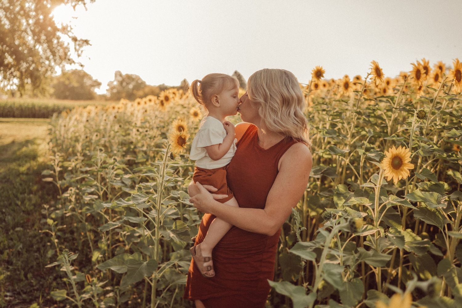 Mother and child share a loving moment in a field of tall sunflowers at sunset.