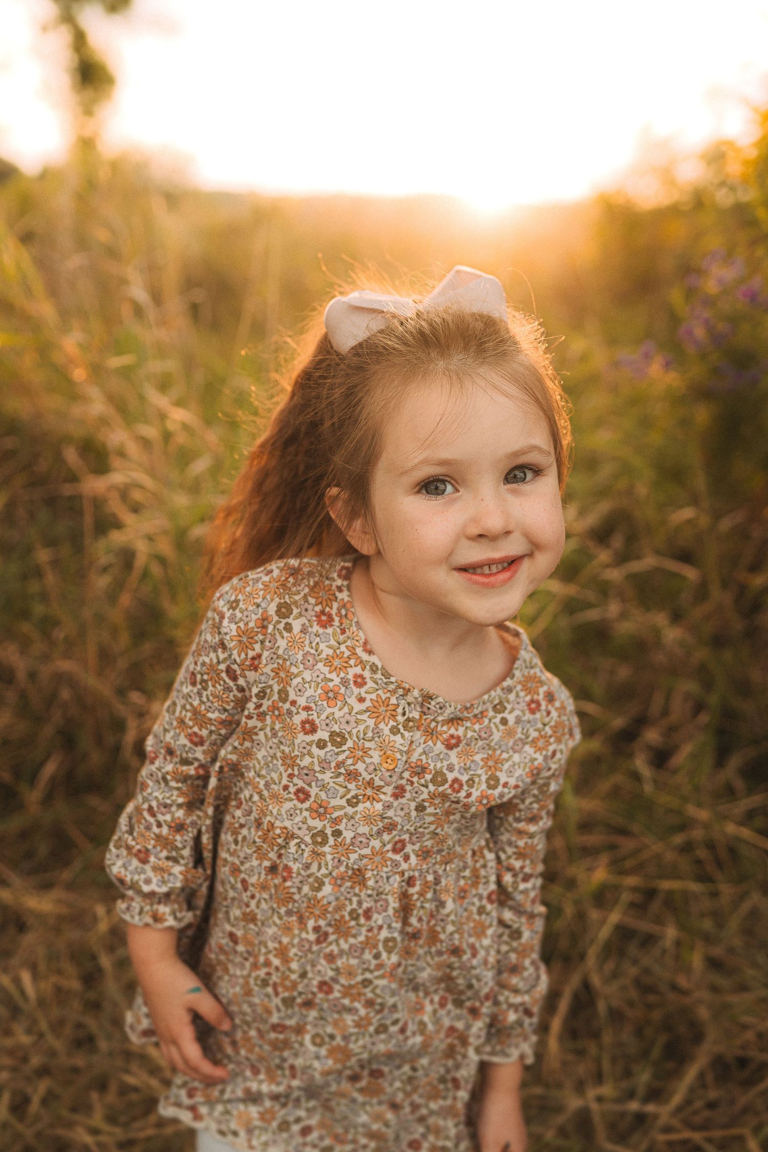 Child in floral dress smiling in a meadow at sunset.