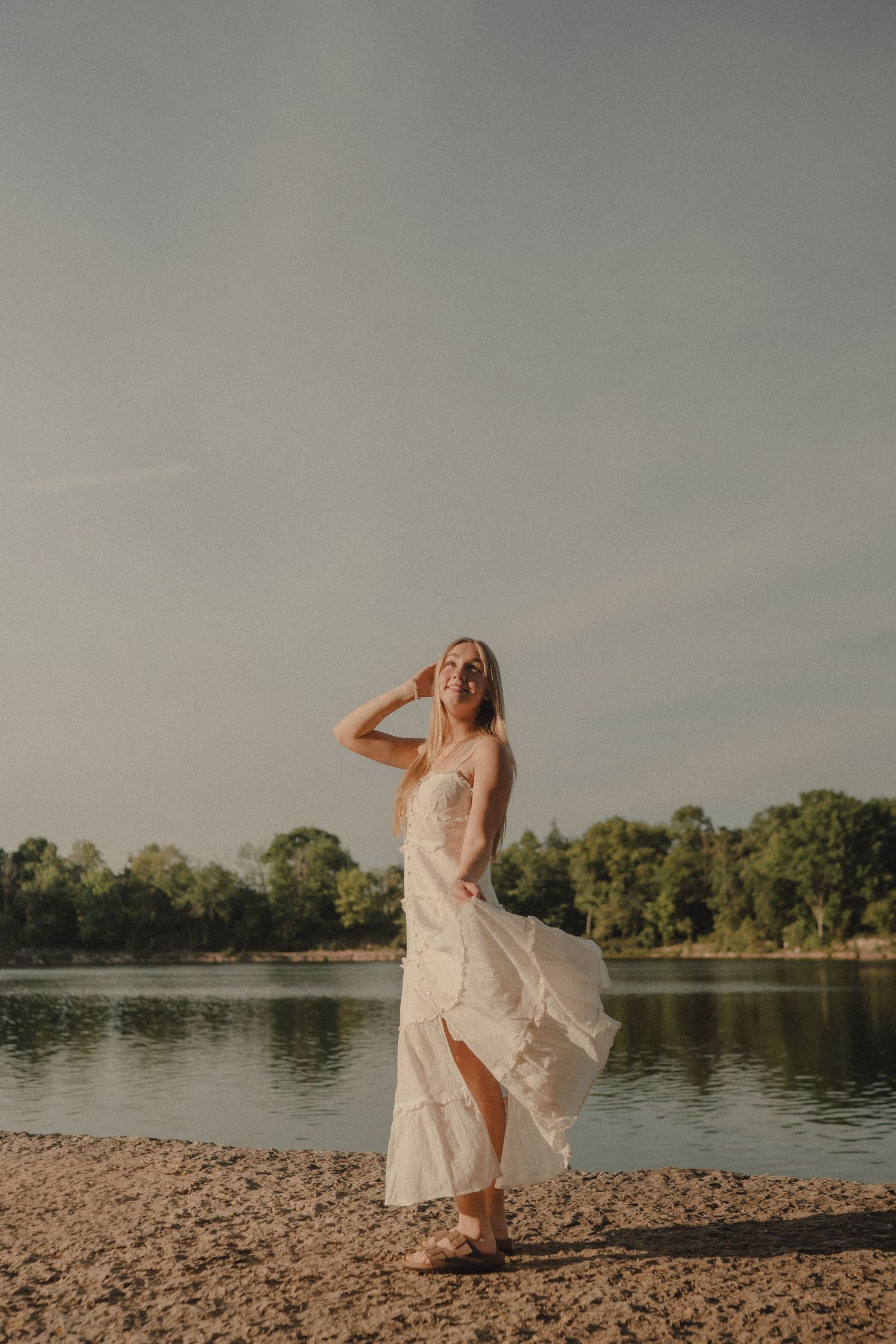 Woman in white lace dress standing on sandy lakeside beach with trees and sky in background.