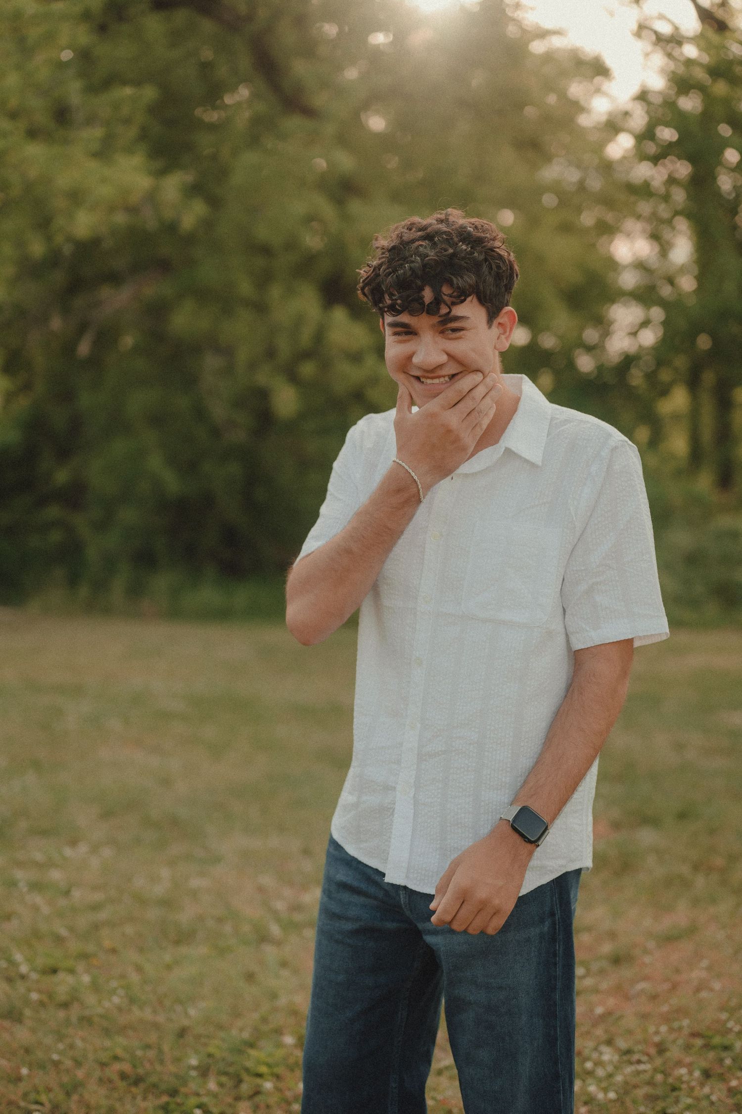Young man with curly hair in white shirt and jeans smiling outdoors in a sunlit green field.