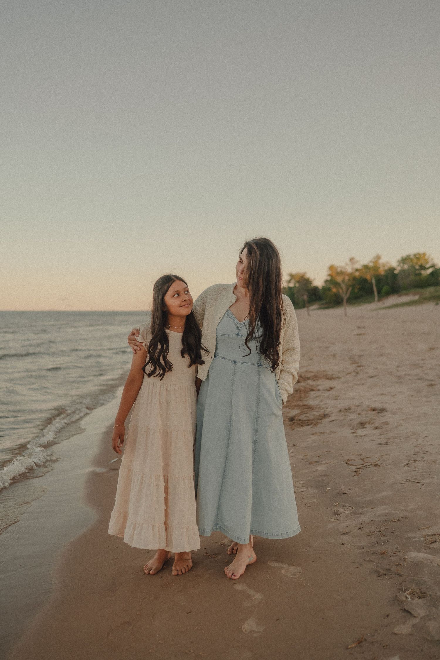 Mother and daughter in flowing dresses walking along sandy beach shore at golden sunset together.