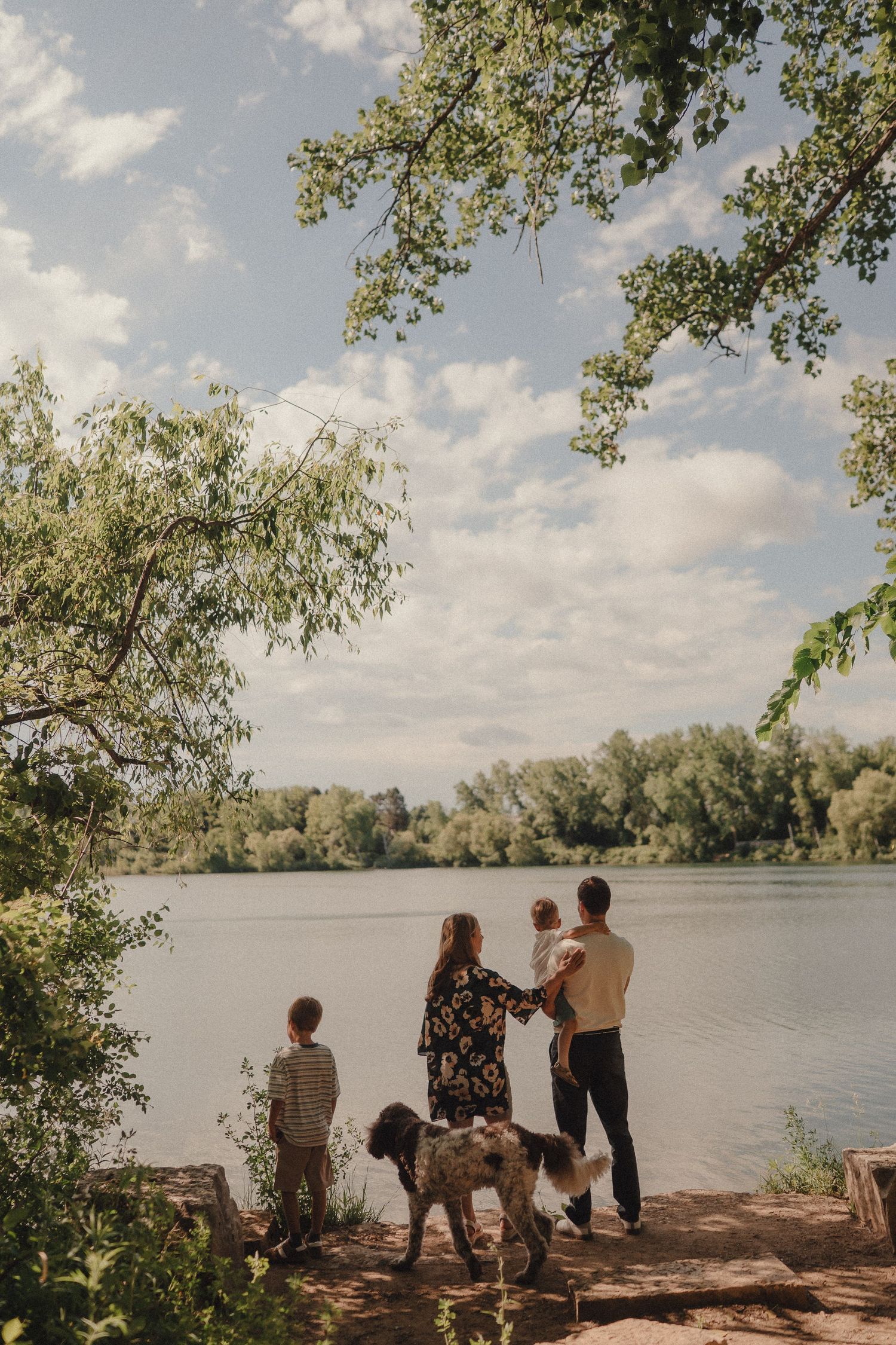 Family with two kids and a dog standing by a scenic lake surrounded by lush green trees.