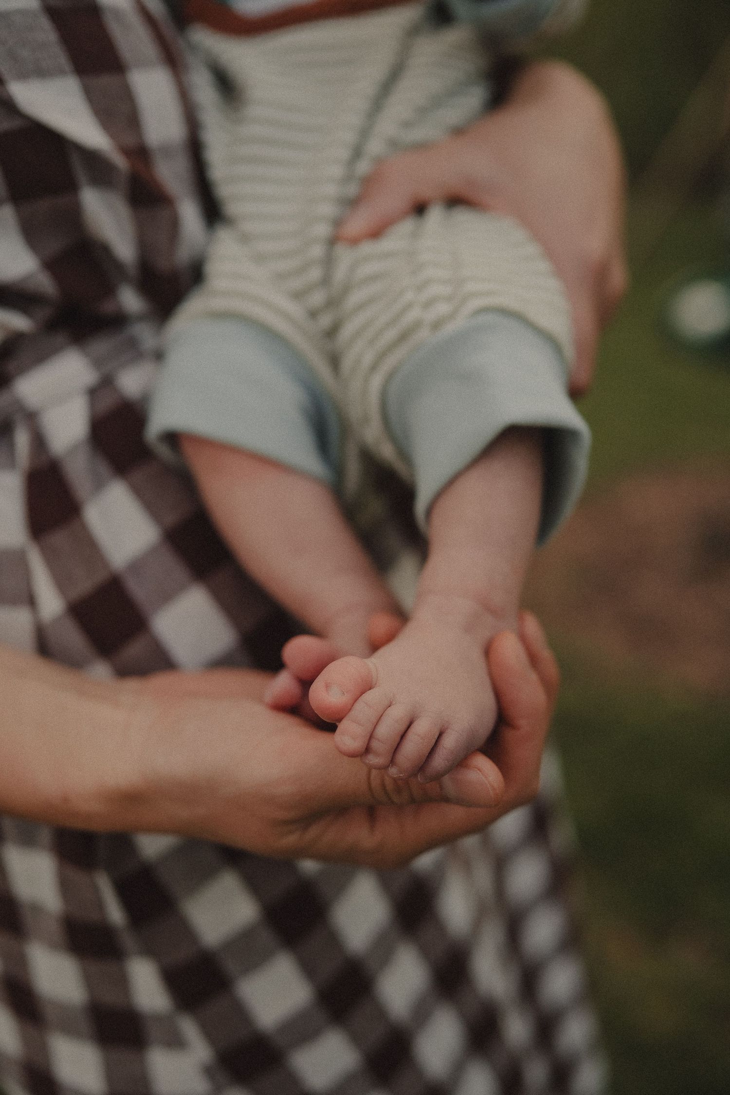 A parent cradles a newborn baby's tiny feet in their hands while wearing a plaid dress outdoors.