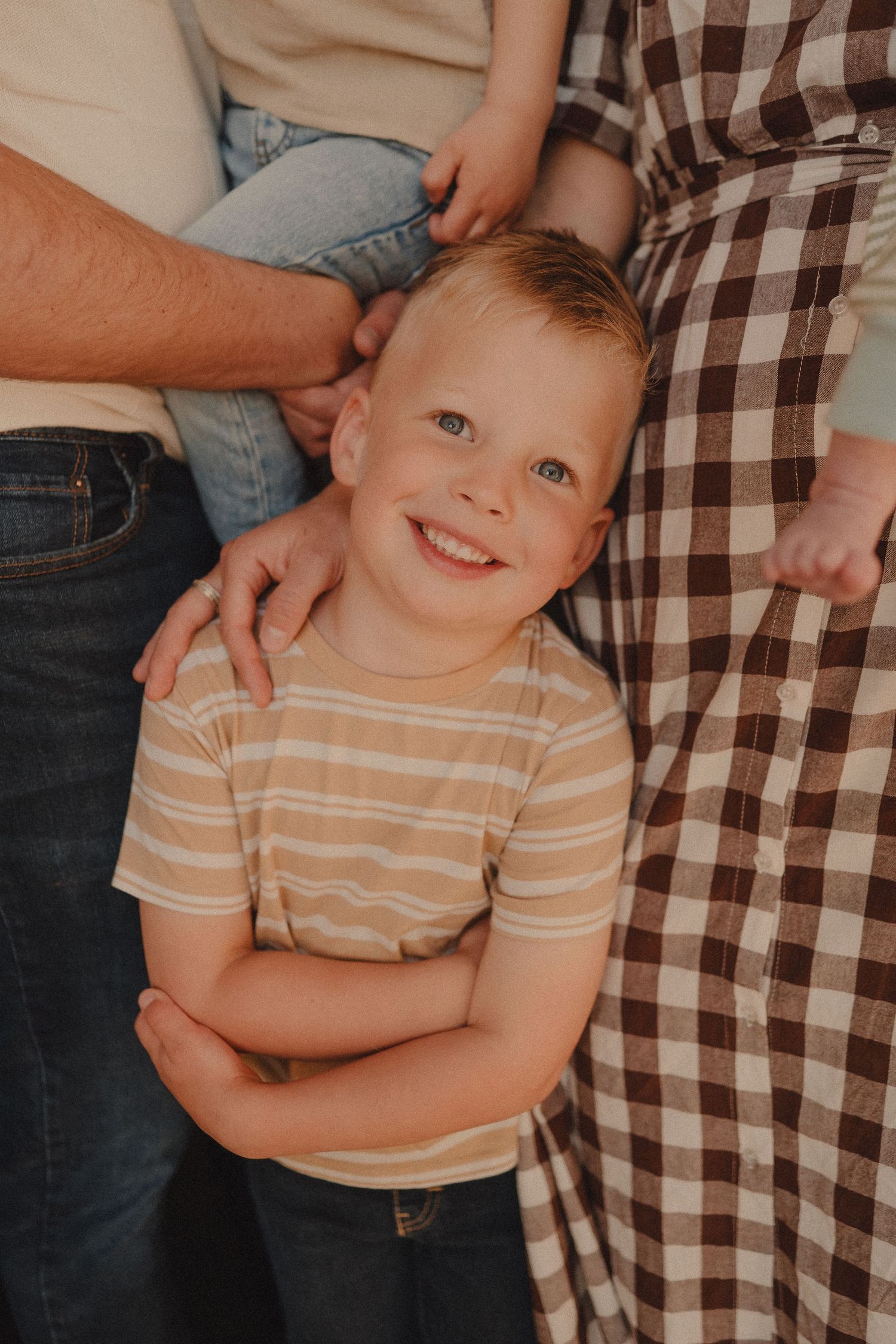 Smiling blonde boy with arms crossed standing between parents in warm outdoor light.