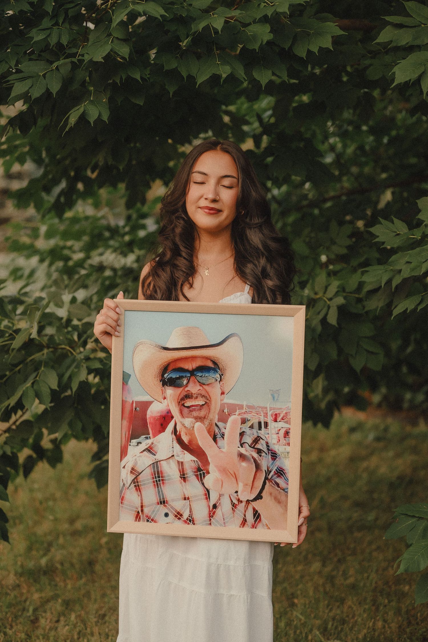 Young woman in white dress holding framed photo of man in cowboy hat outdoors.