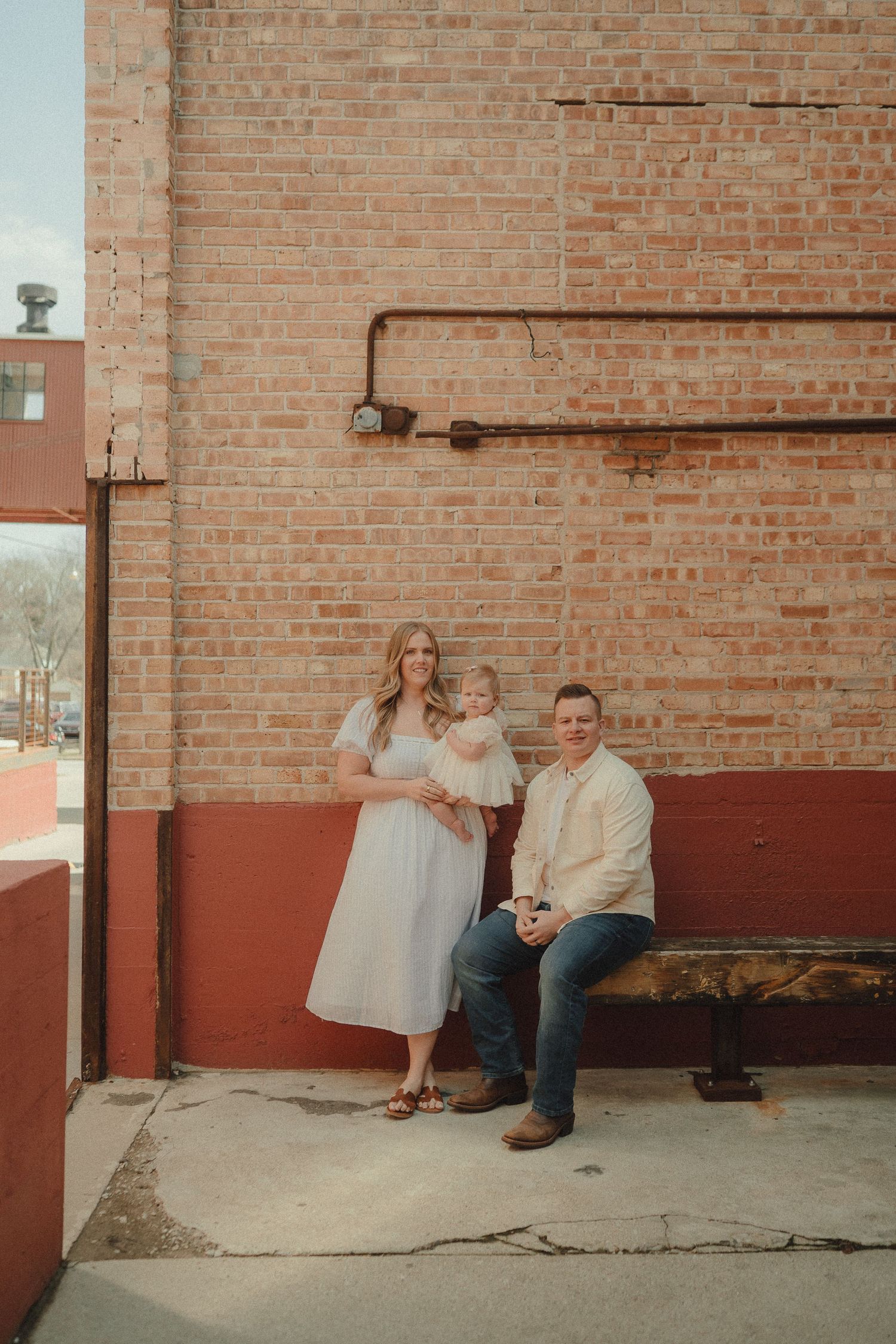 Family of three posing against a rustic brick wall outdoors, mother holding baby girl.