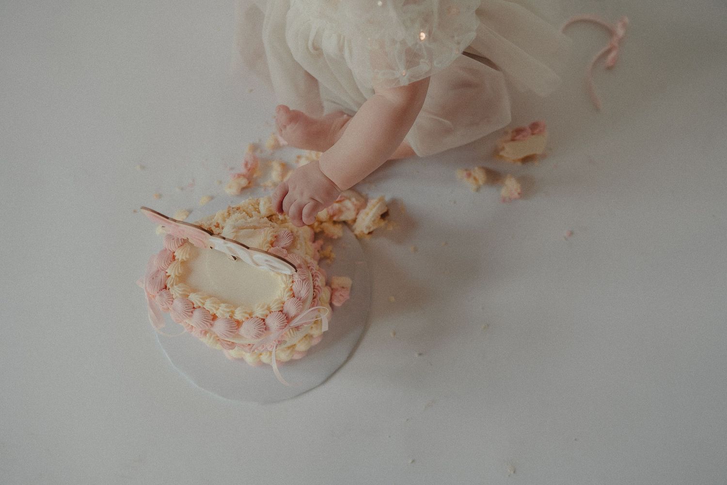 A baby in a white tutu smashes her first birthday cake, grabbing pieces with tiny hands on a white surface.