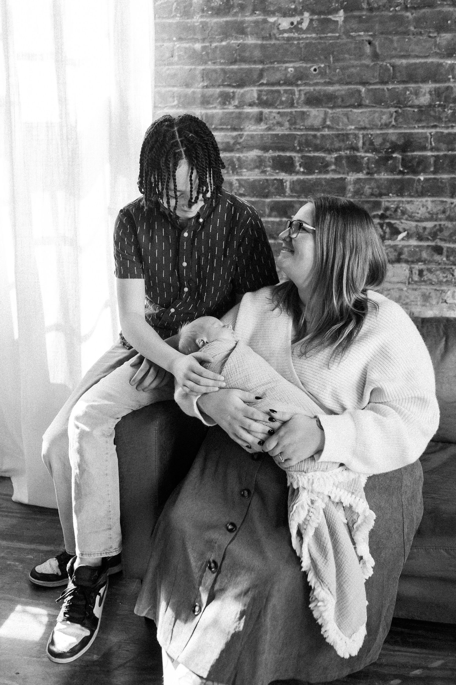 Black and white photo of a mother holding a newborn while an older child with dreadlocks sits beside her on a chair.