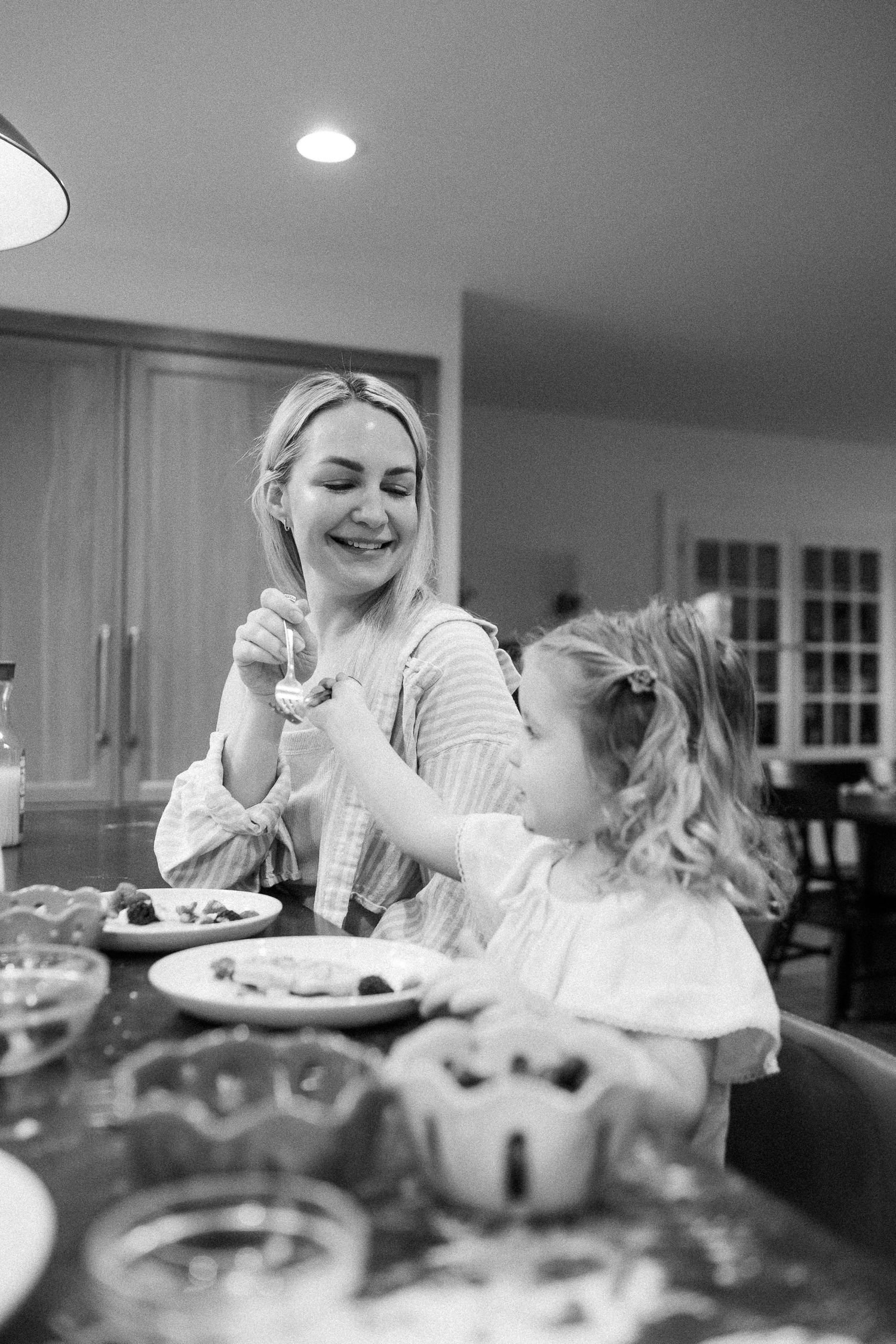 Black and white photo of a mother smiling as her young daughter feeds her a treat at the kitchen counter.