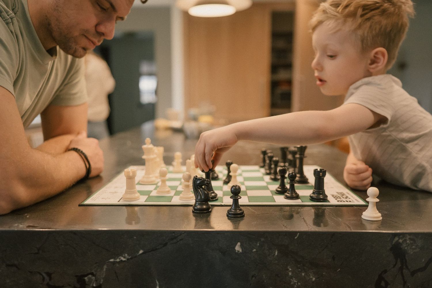 A father and young son play chess together at a dark marble kitchen counter.