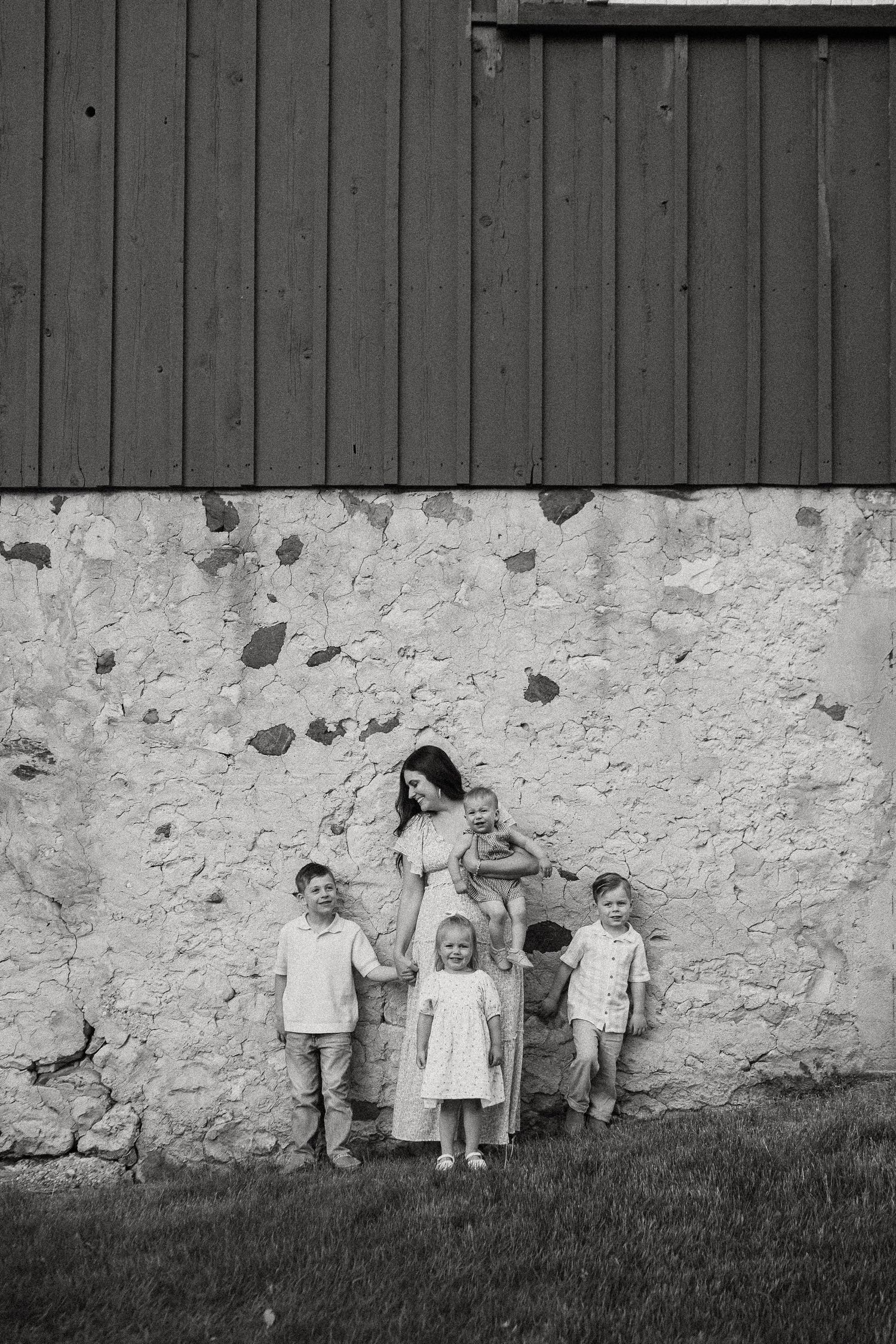 Black and white photo of a mother with four young children posed against a rustic stone barn wall.