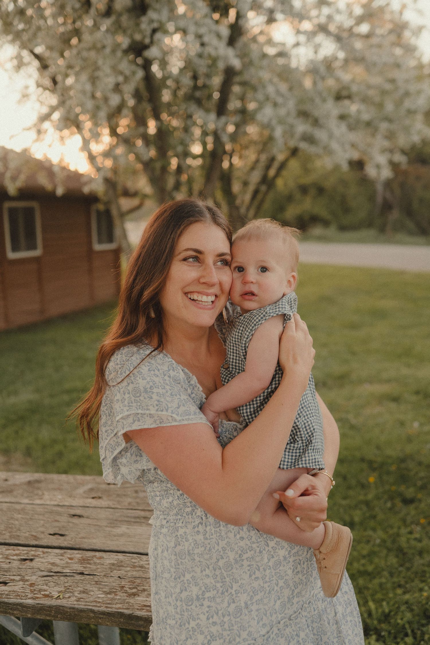 Happy mother in floral dress holding baby in gingham outfit, smiling near blooming white cherry tree at sunset.