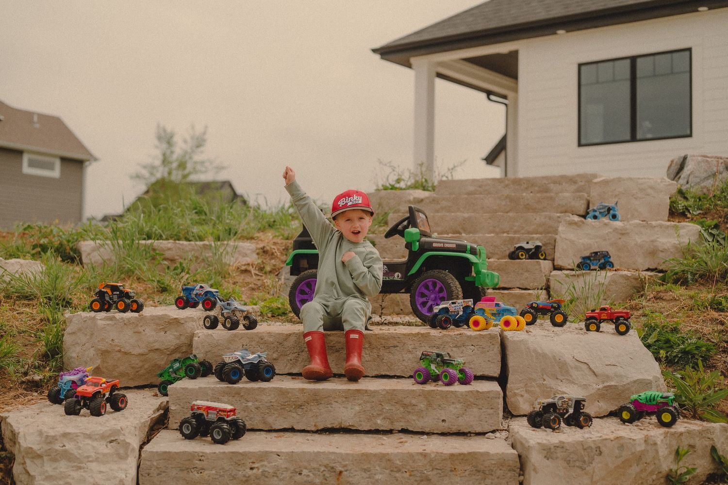 Happy toddler surrounded by monster truck toys on stone steps outside new home.