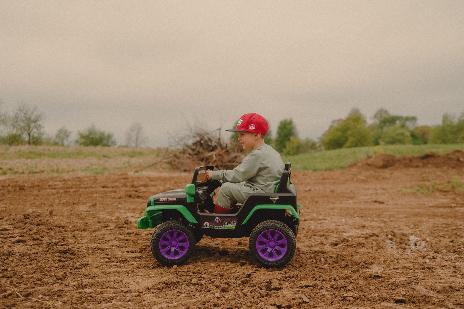 Young child wearing red hard hat driving green and purple toy jeep on dirt field outdoors.