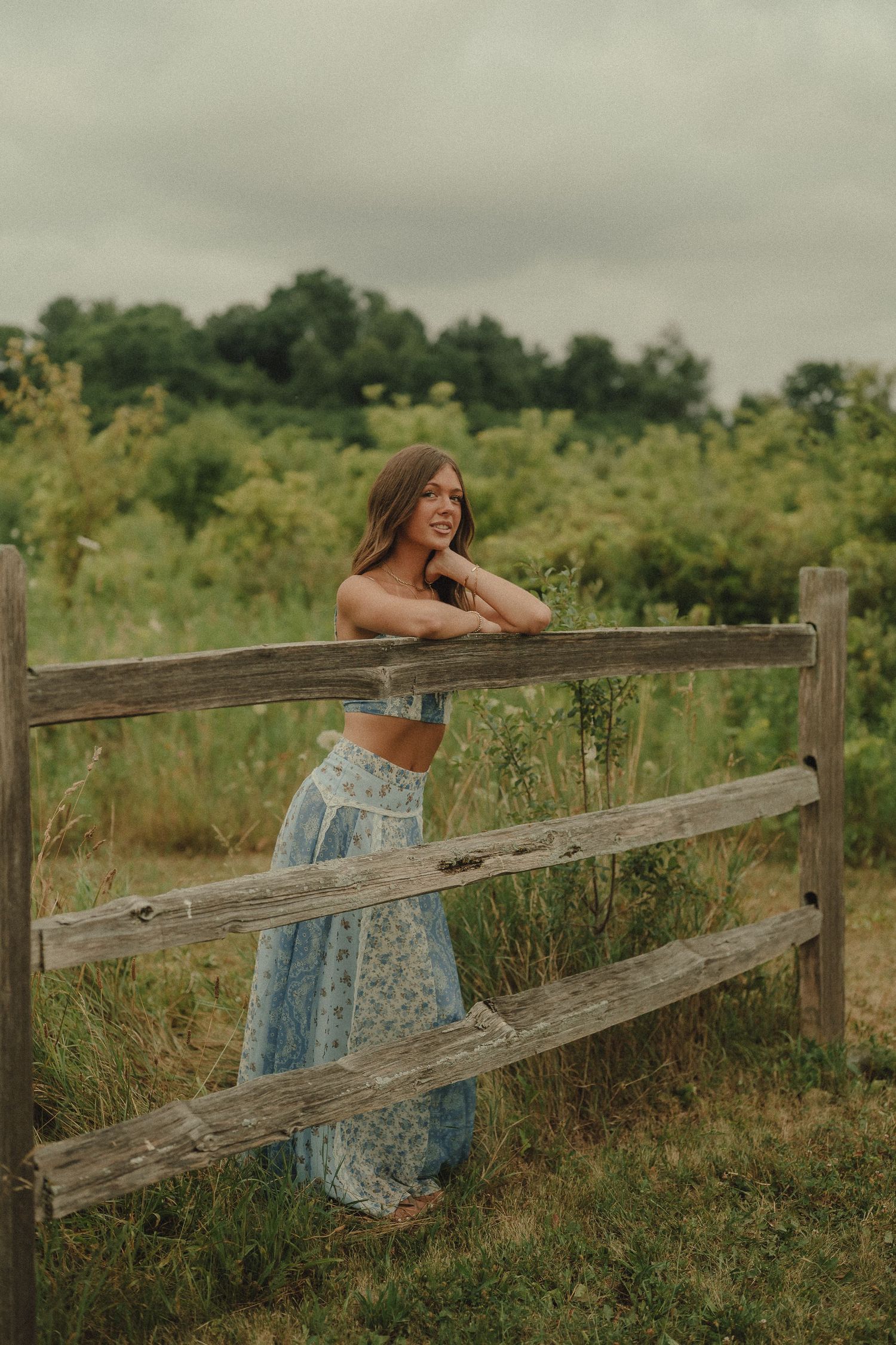 Young woman in blue floral crop top and skirt leaning on a rustic wooden fence in a lush green field.