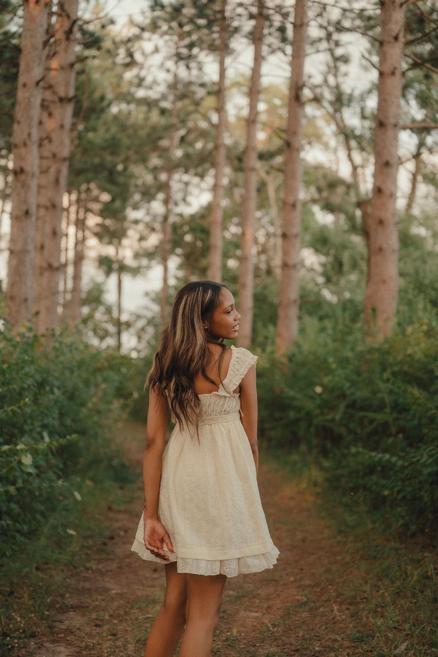 Young woman in white dress standing on forest path surrounded by tall trees.