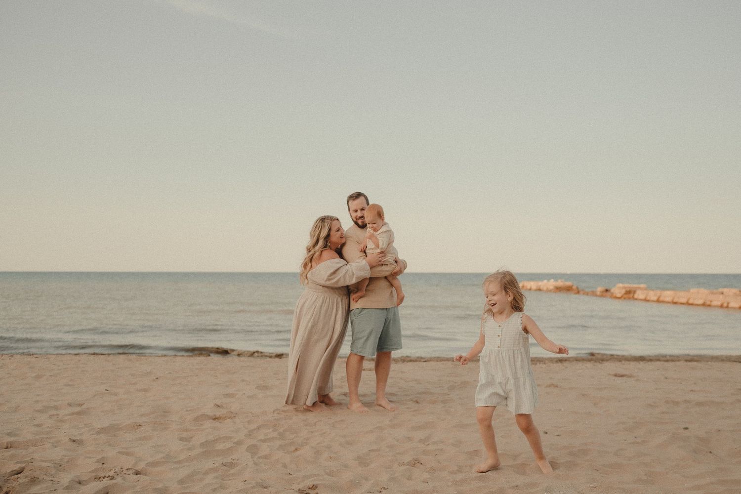 Happy family of four on sandy beach at golden hour, father holding baby while girl dances nearby.