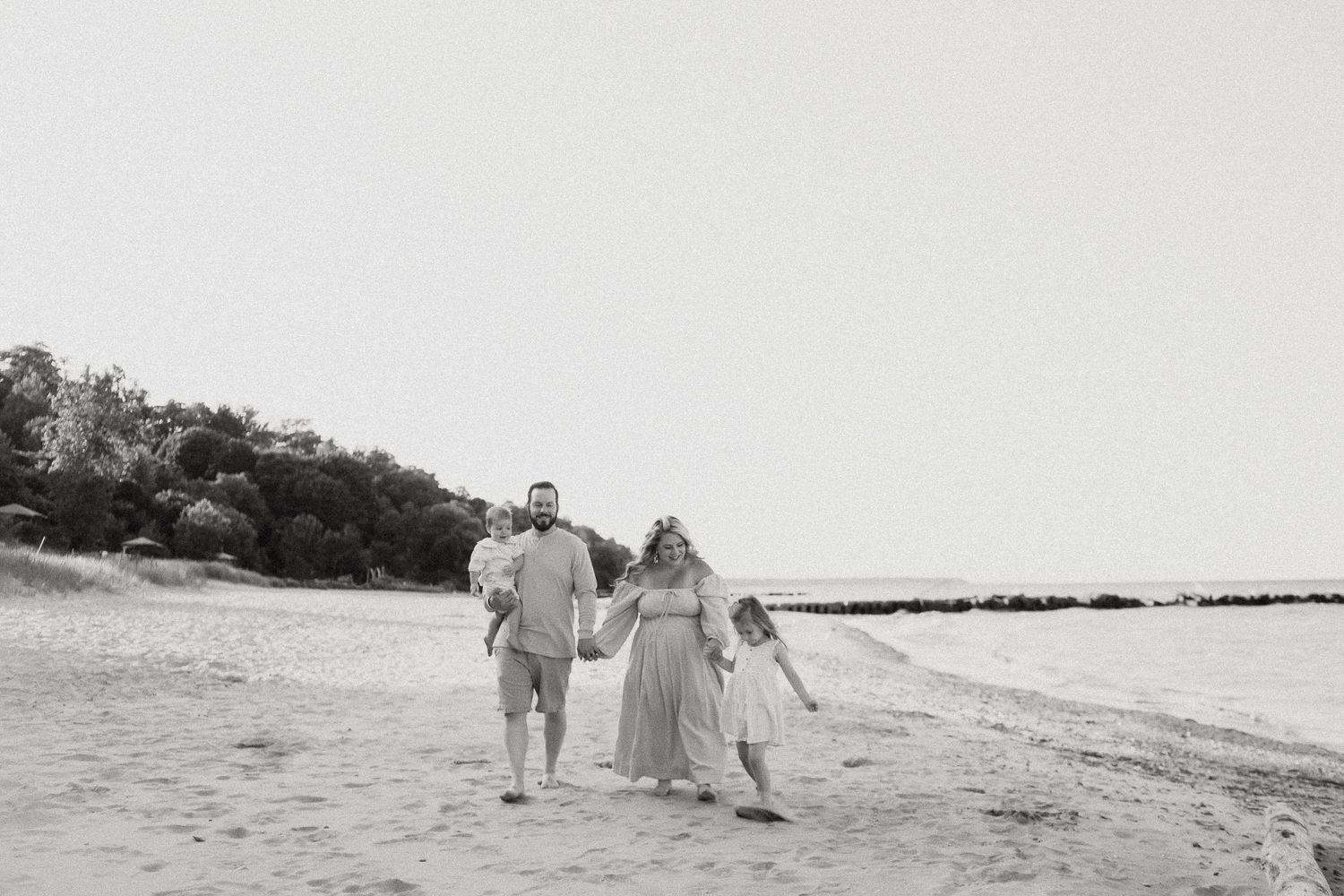 Couple holding hands walking on beach in black and white photo with trees and water in background.