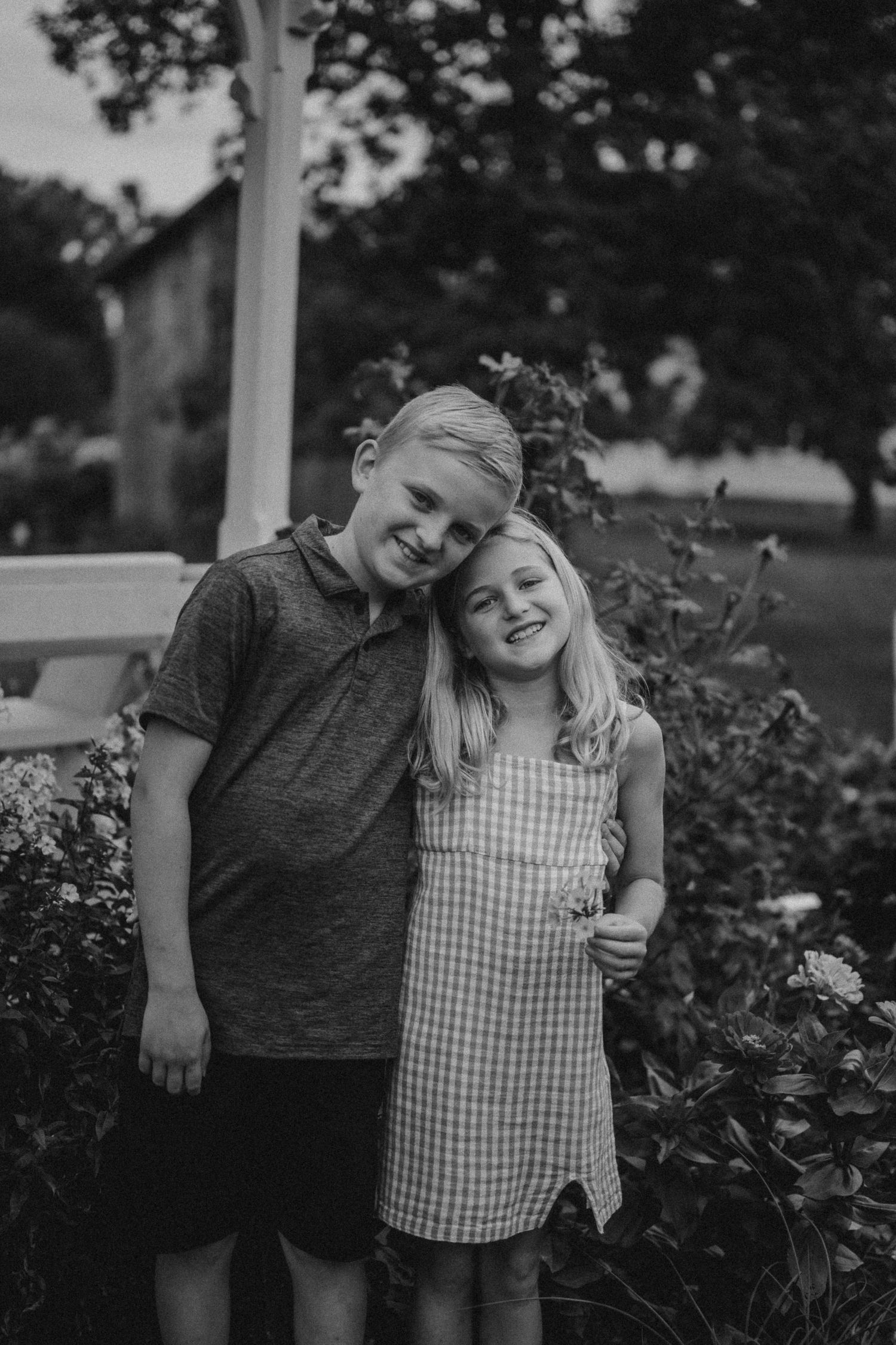 Black and white portrait of two children embracing by a flowering garden.