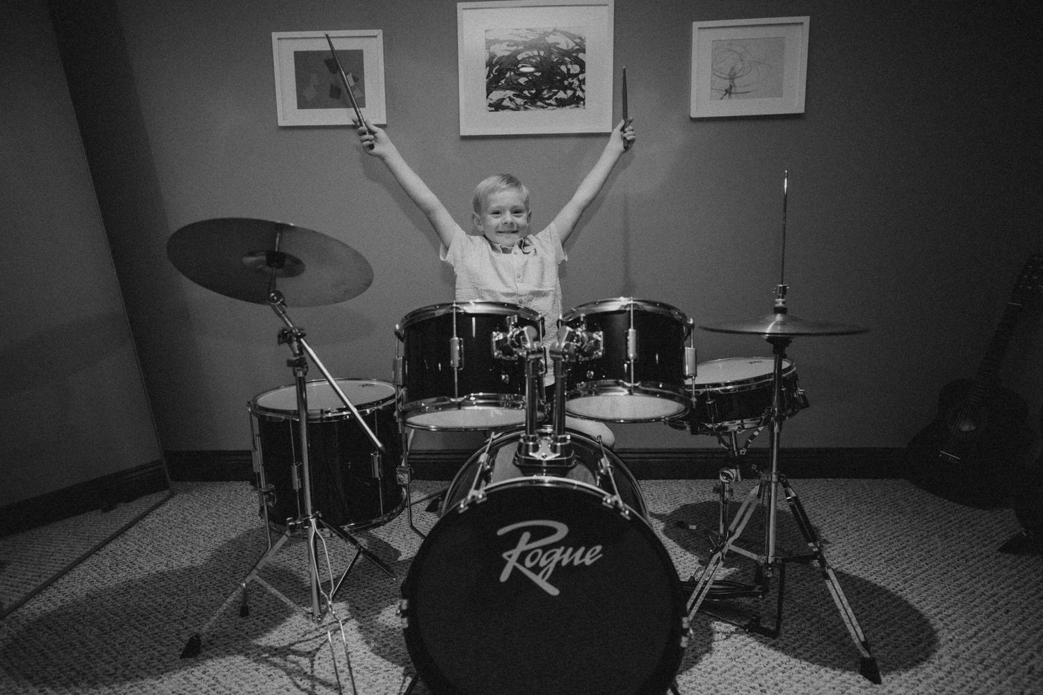 A child enthusiastically playing drums with raised arms in a black and white photo.
