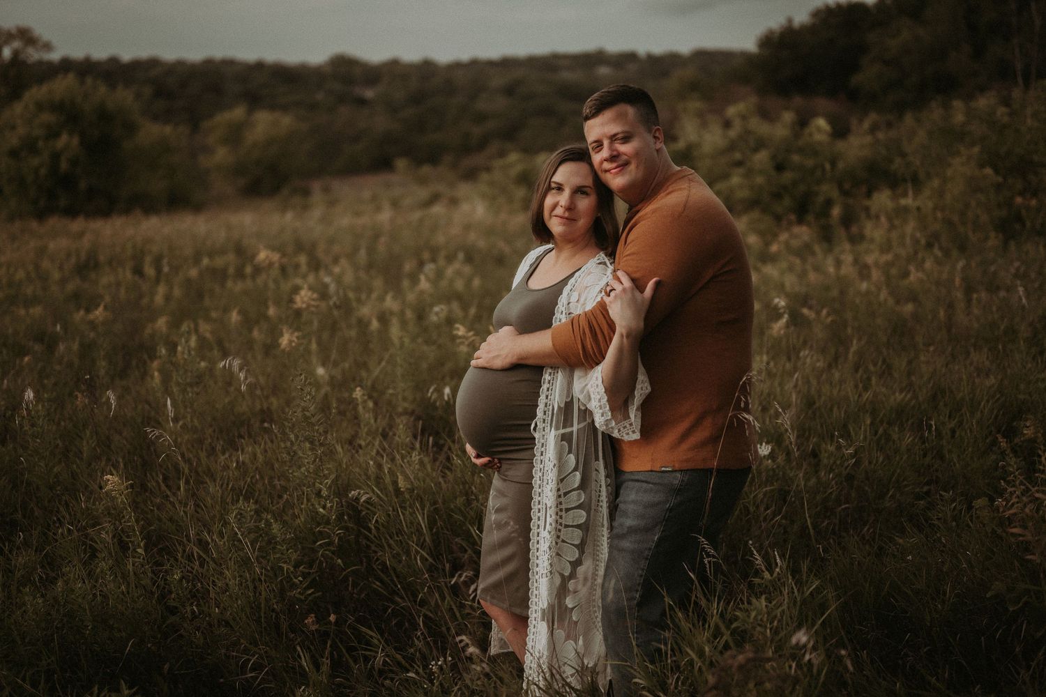 Expecting couple shares an intimate moment in a rustic field with moody sky backdrop.